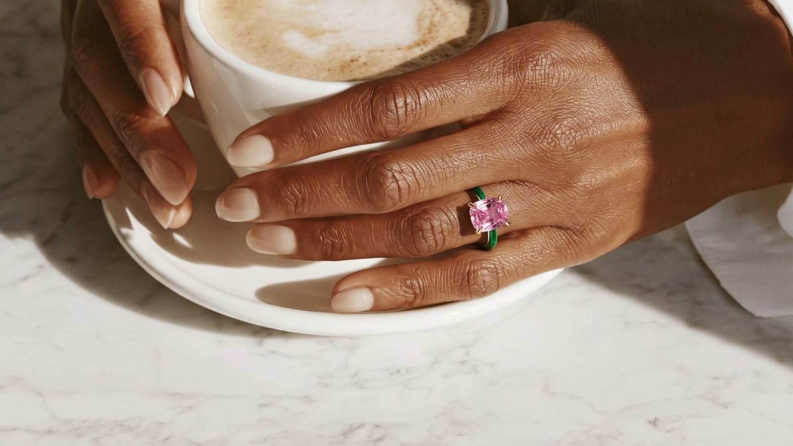 Hand with a green enamel ring and pink gemstone holding a coffee cup