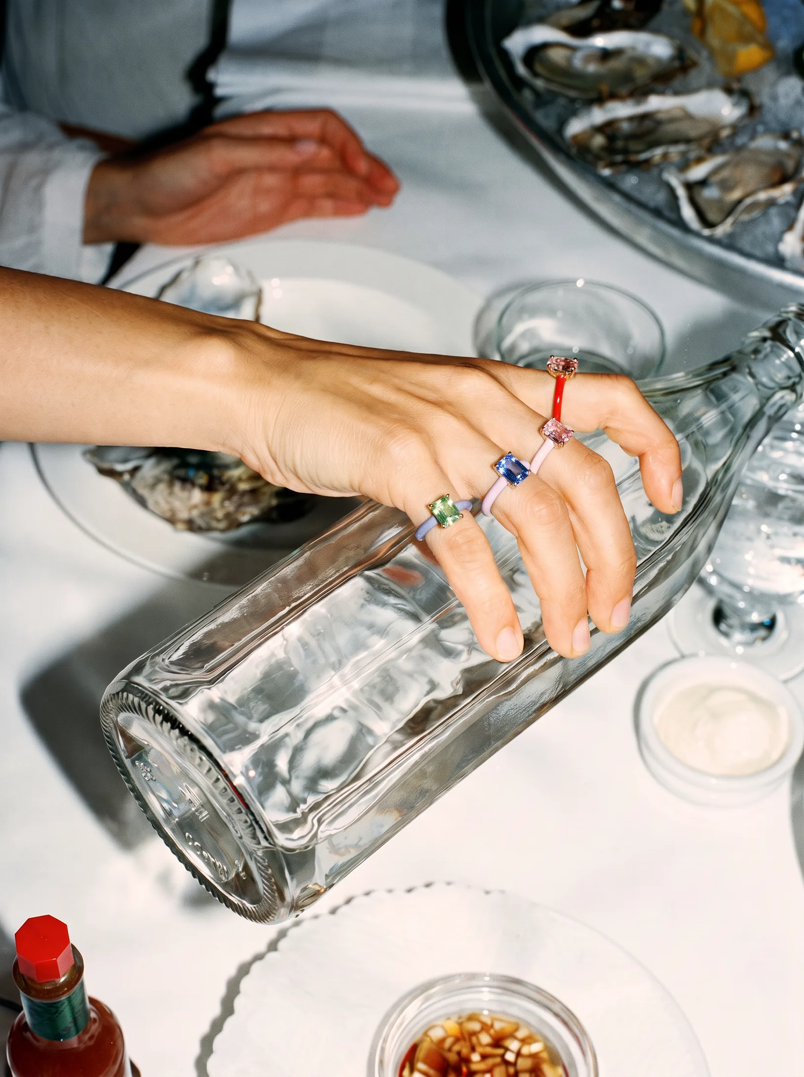 Hand wearing colorful gemstone and enamel rings holding a glass water bottle